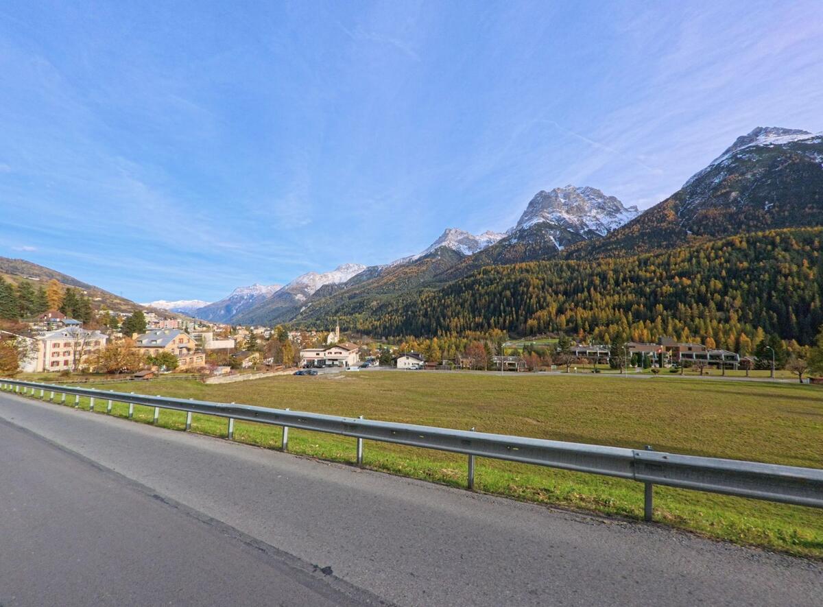 Über offene Wiesen hinweg zeigt sich in Scuol (7550, Kanton Graubünden) ein Dorfteil mit dicht stehenden Wohnhäusern am Hang vor herbstlich gefärbten Wäldern.