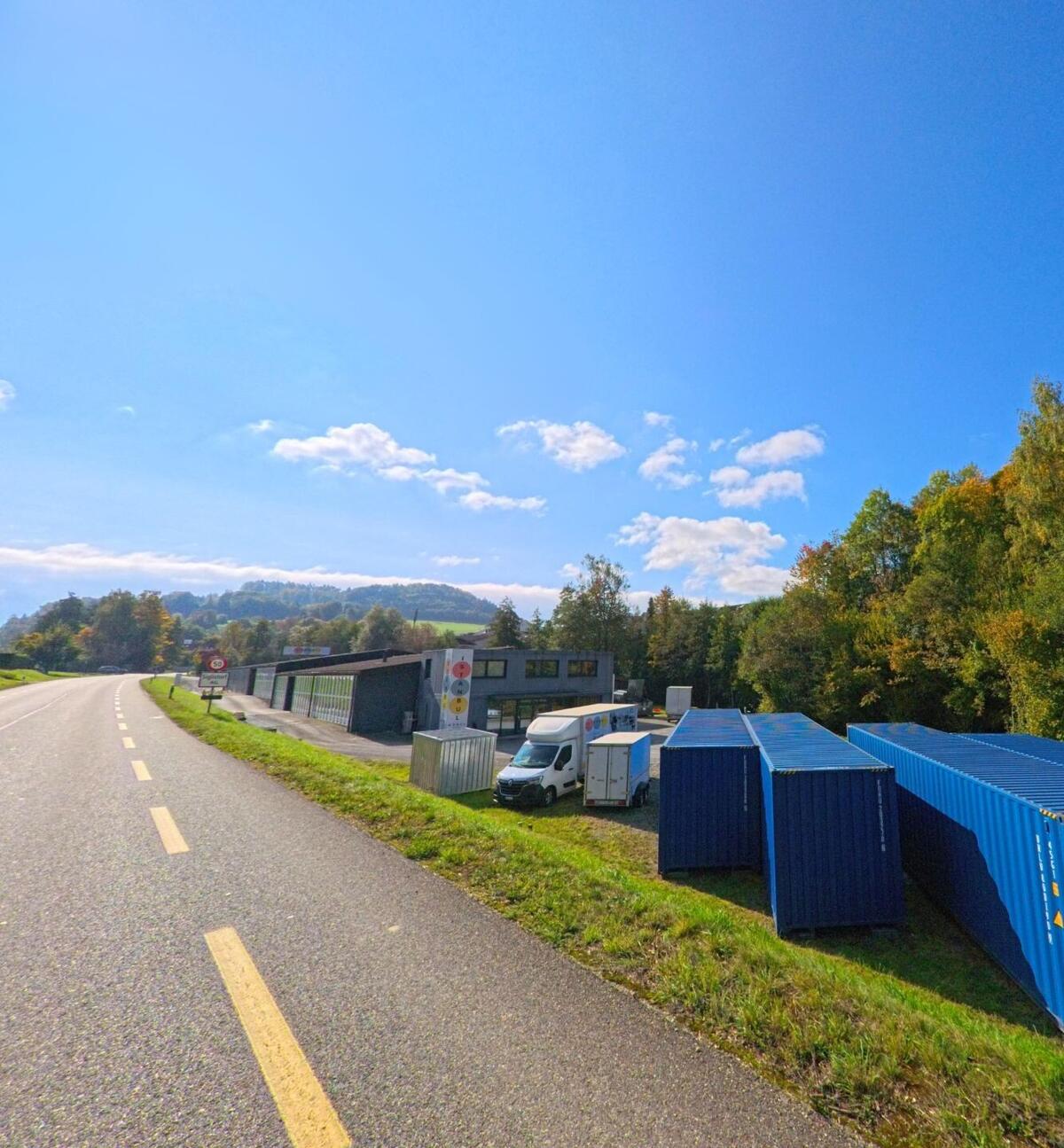 Neben einer leicht geschwungenen Landstrasse in Siglistorf (5462, Kanton Aargau) stehen Industriegebaeude, Transporter und mehrere blaue Container vor einem bewaldeten Huegel.