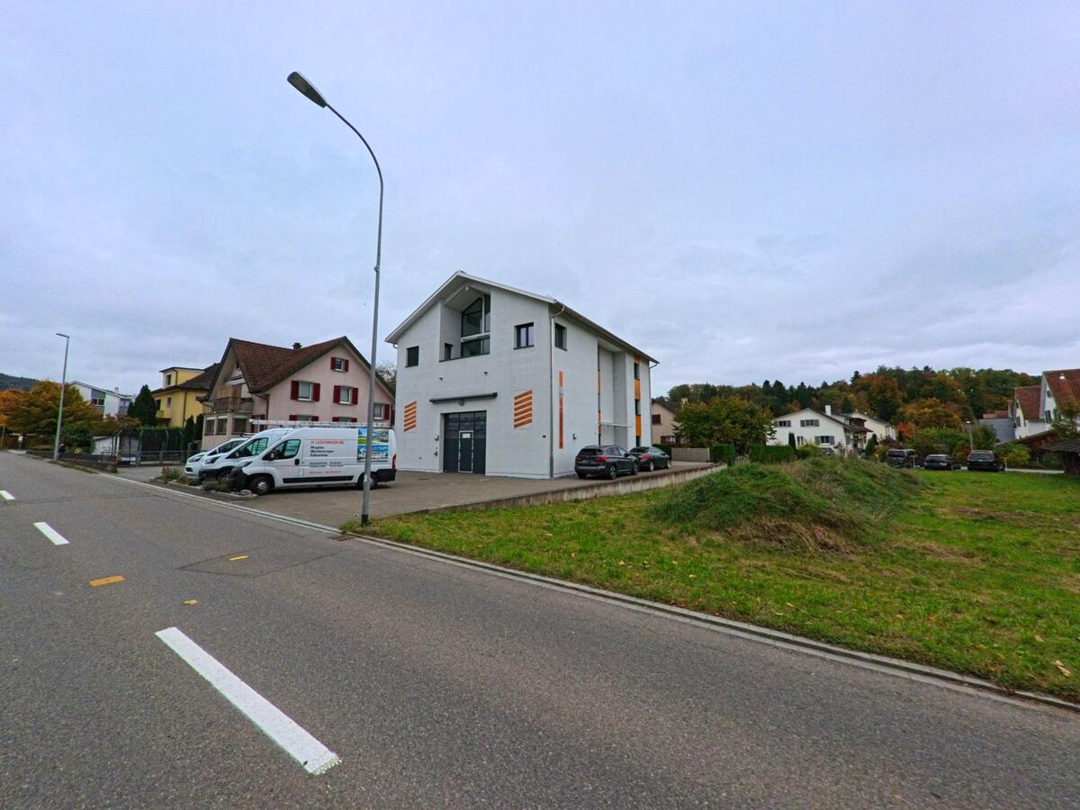 An einer Quartierstrasse mit angrenzender Grünfläche steht ein modernes Gewerbe- oder Wohngebäude zwischen Einfamilienhäusern in Aadorf (8355, Kanton Thurgau).