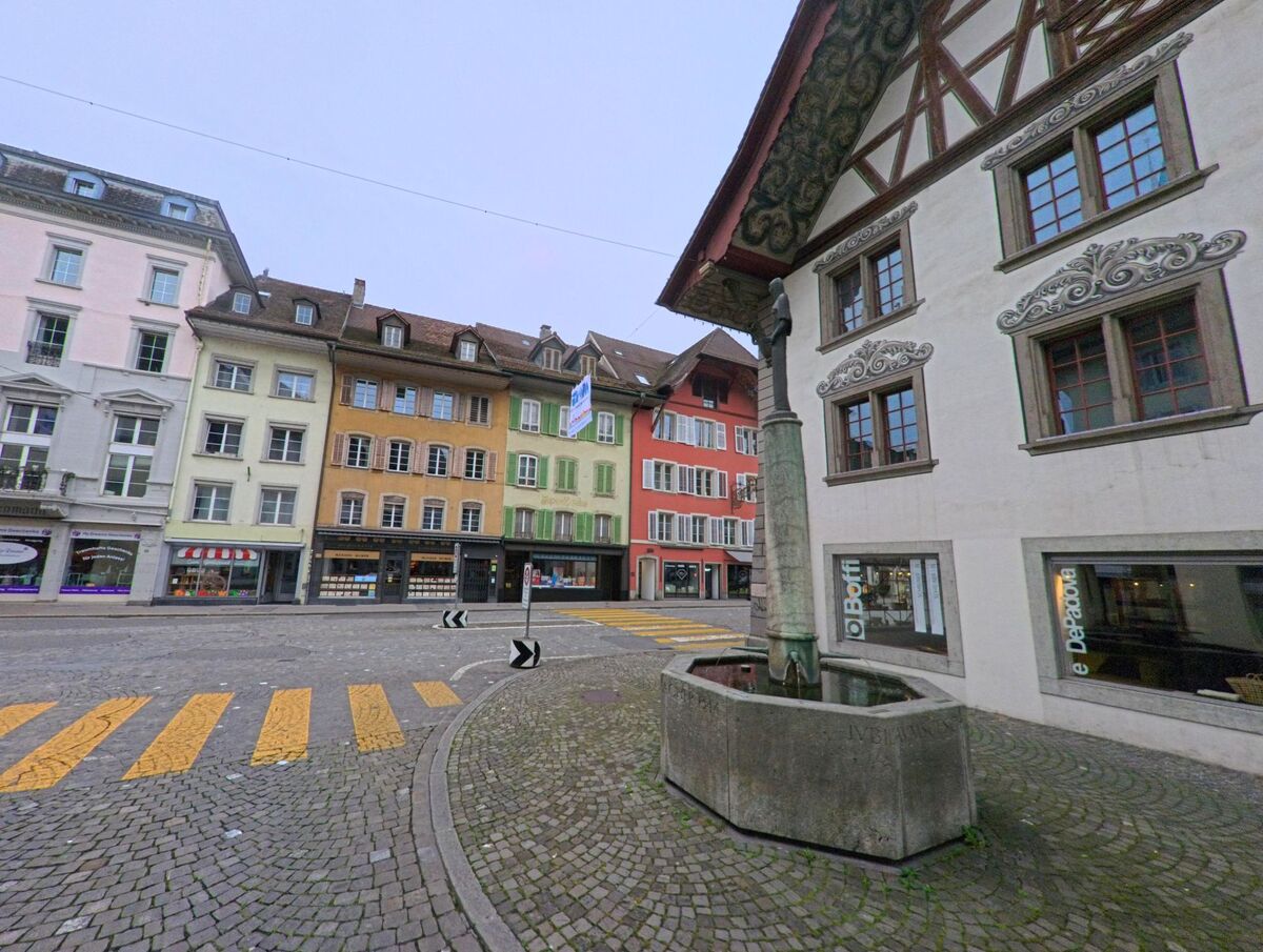 In Aarau (5000, Kanton Aargau) säumen farbenfrohe historische Häuser den zentralen Platz mit Kopfsteinpflaster, im Vordergrund steht ein Brunnen mit steinerner Säule.