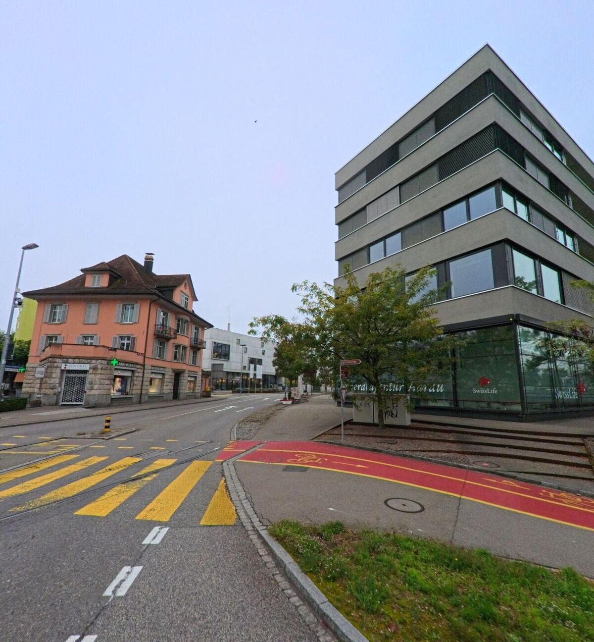 An einer Strassenecke in Aarau (5000, Kanton Aargau) treffen ein modernes Bürogebäude und ein älteres Wohnhaus aufeinander, getrennt durch einen markierten Fussgängerstreifen
