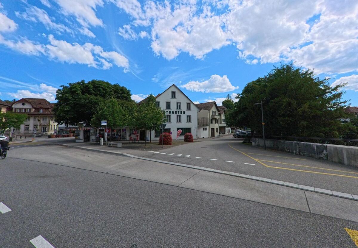 In Aarburg (4663, Kanton Aargau) steht das Gebaeude des Restaurants Stadtgarten an einer Strasseneinmuendung mit angrenzendem Kiosk und schattigen Baeumen.