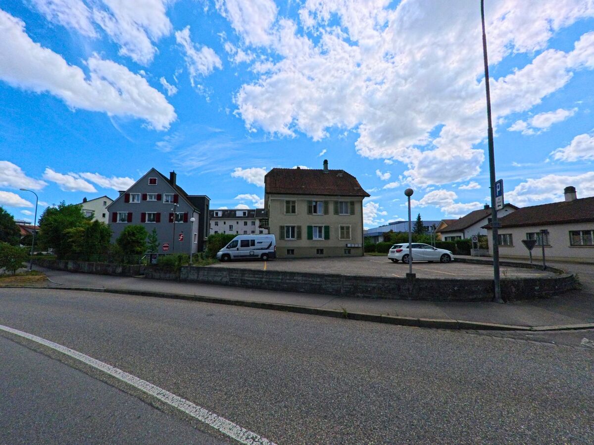 Mehrere Wohnhaeuser mit Vorplaetzen und ein kleiner Parkplatz praegen das ruhige Quartierbild in Aarburg (4663, Kanton Aargau) an einer leicht gekruemmten Strasse.