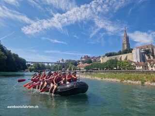 Eine Gruppe von Personen paddelt bei Aarebootsfahrten CH in Steffisburg (3613, Kanton Bern) in einem Schlauchboot auf der Aare mit Blick auf die Berner Altstadt.