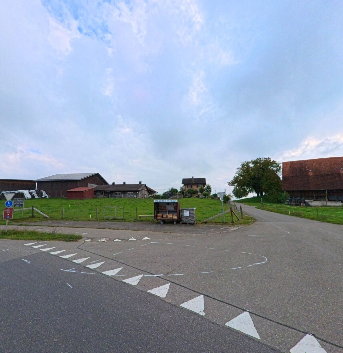 Am Dorfrand von Abtwil (5646, Kanton Aargau) liegen Bauernhoefe und Wiesen entlang einer Abzweigung mit einem kleinen Verkaufsstand.