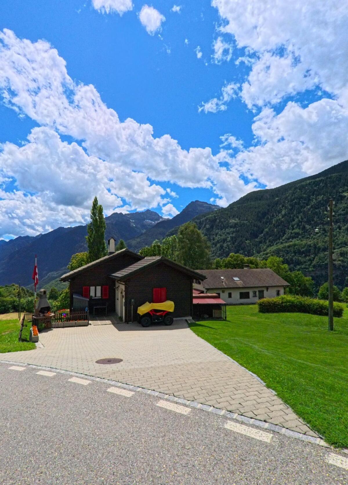 Ein einzelnes Chalet mit roten Fensterläden steht in Faido (6746, Kanton Tessin) auf einer grünen Wiese mit Blick auf bewaldete Berghänge und den blauen Himmel.