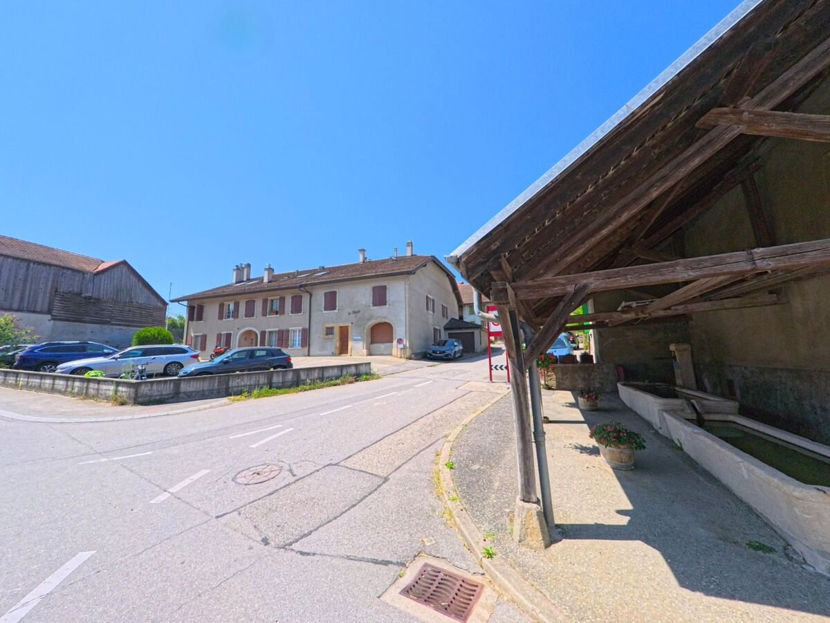 Ein gedeckter Dorfbrunnen mit langen Wasserbecken steht in Aclens (1123, Kanton Waadt) neben traditionellen Wohnhaeusern und einer schmalen Strasse.