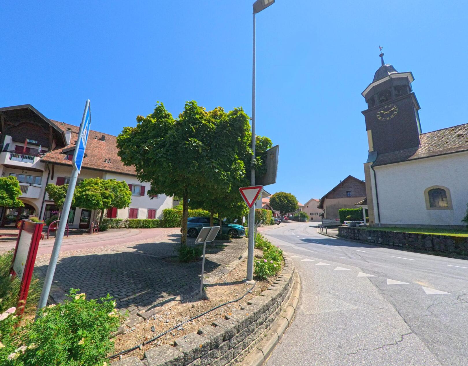 Im Zentrum von Aclens (1123, Kanton Waadt) steht die Kirche mit ihrem markanten Turm und einer grossen Uhr, daneben fuehrt die Hauptstrasse durch das Dorf.