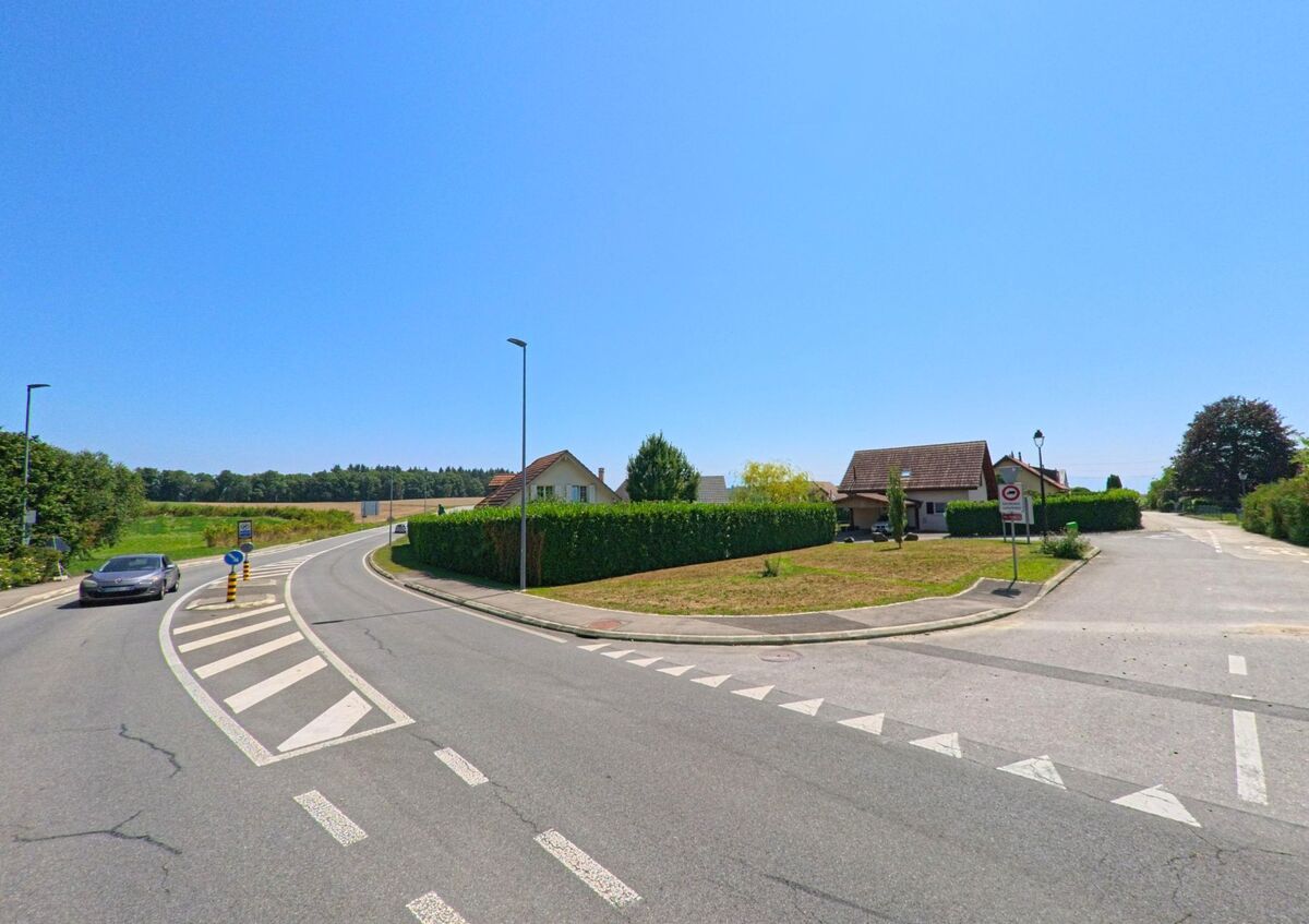 Am Rand von Aclens (1123, Kanton Waadt) fuehrt eine Hauptstrasse an Einfamilienhaeusern mit gepflegten Hecken vorbei, waehrend Felder im Hintergrund liegen.