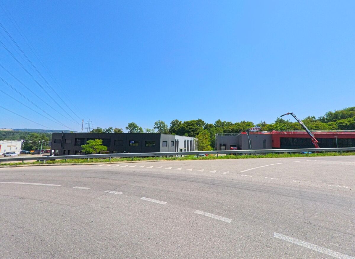 Am Rand von Aclens (1123, Kanton Waadt) stehen moderne Industriegebaeude, darunter eine Halle mit der Aufschrift „Eclair“, neben einer breiten Strasse.