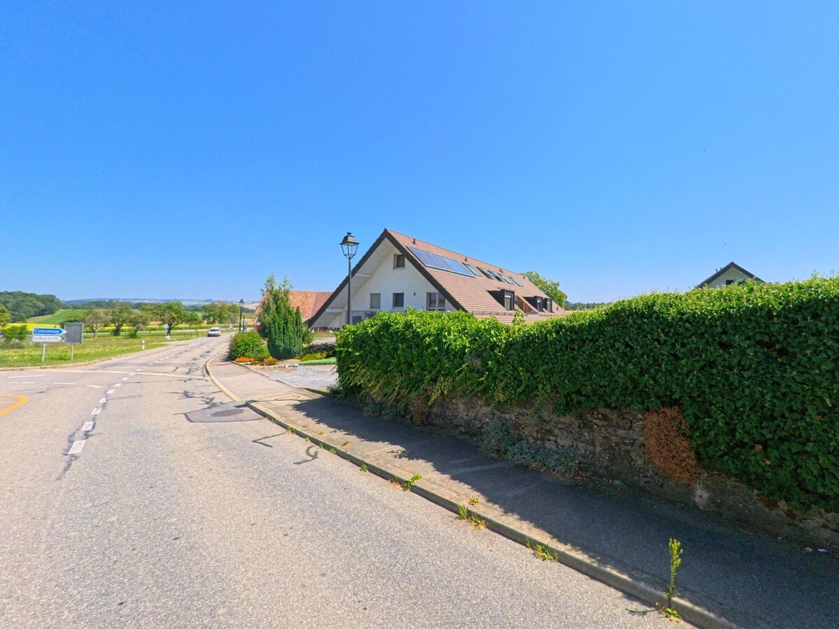 In Aclens (1123, Kanton Waadt) verläuft eine Landstrasse mit Blick auf Felder und Obstbaeume, waehrend ein Wohnhaus mit Solarpanelen am Ortsrand steht.