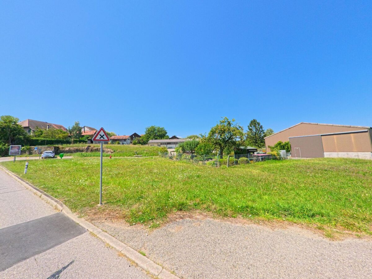 Eine Nebenstrasse in Aclens (1123, Kanton Waadt) fuehrt an einer Gruenflaeche mit Verkehrsschild vorbei, waehrend Wohnhaeuser und eine grosse Halle im Hintergrund sichtbar sind.