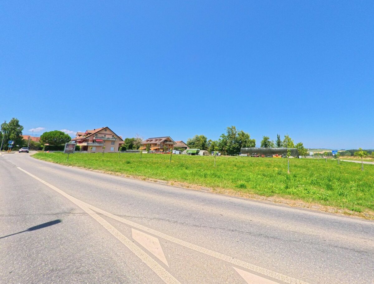 An einer breiten Strasse in Aclens (1123, Kanton Waadt) liegen Wiesen, Wohnhaeuser und Gewaechshaeuser, im Hintergrund breitet sich die Landschaft aus.