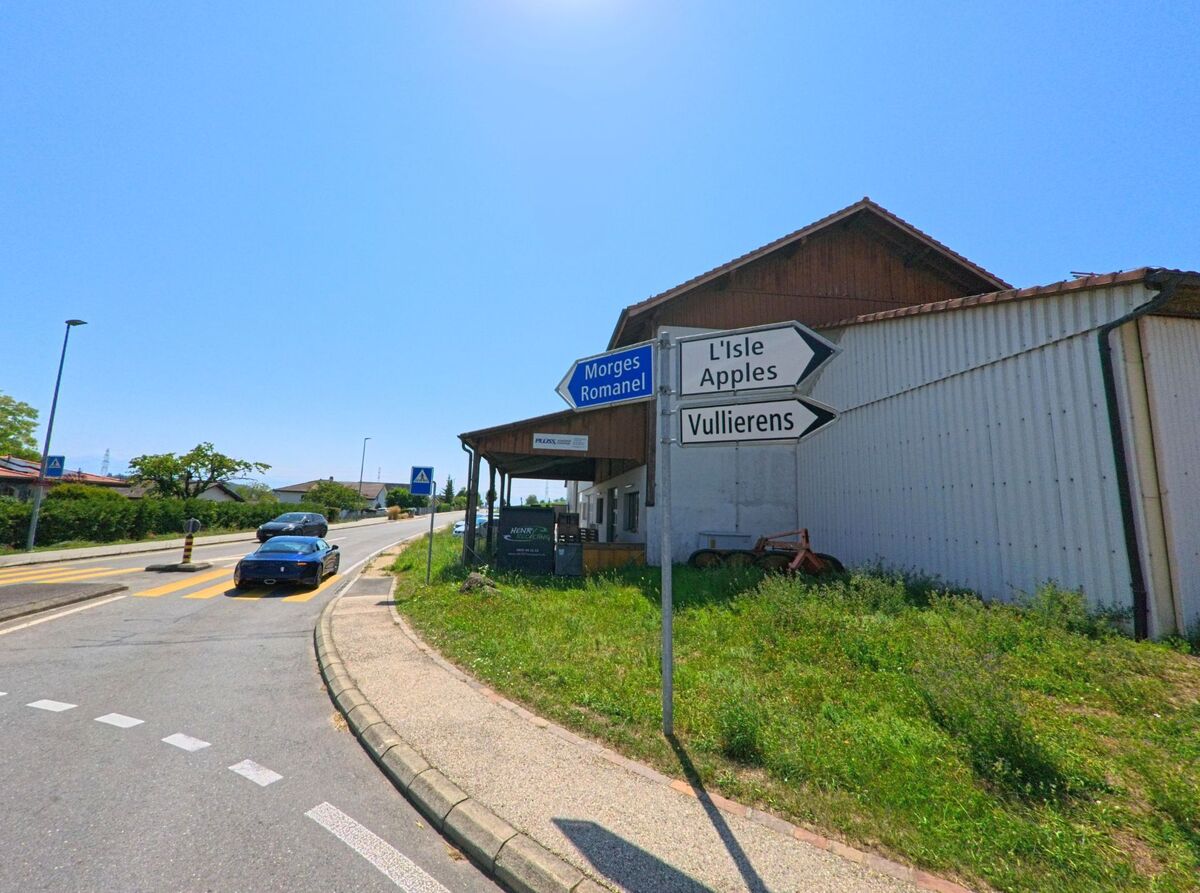 Ein Wegweiser in Aclens (1123, Kanton Waadt) zeigt die Richtungen nach Morges, Romanel, L’Isle, Apples und Vullierens, daneben verlaeuft die Hauptstrasse am Ortsrand.