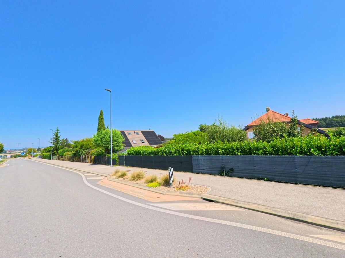 Eine Wohnstrasse in Aclens (1123, Kanton Waadt) fuehrt an Einfamilienhaeusern mit Gaerten und hohen Hecken entlang, im Hintergrund sind Felder sichtbar.