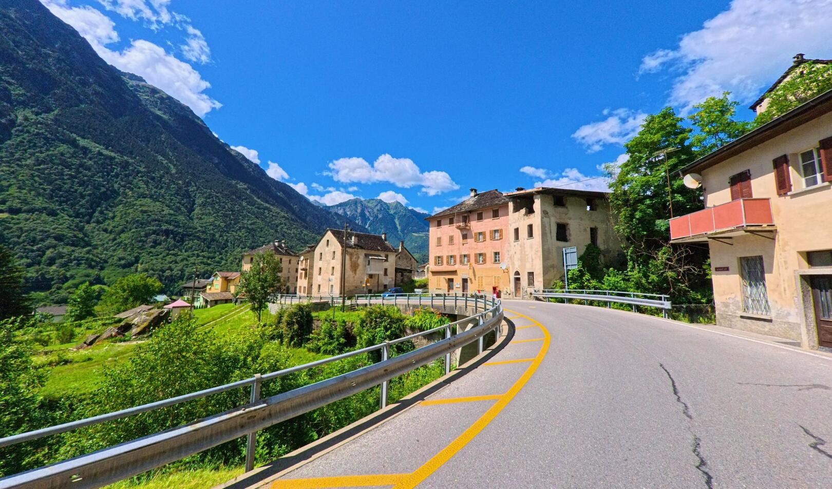 Eine kurvenreiche Strasse fuehrt durch das historische Dorf von Acquarossa (6723, Kanton Tessin) mit alten Haeusern und Blick auf die umliegenden Berge.