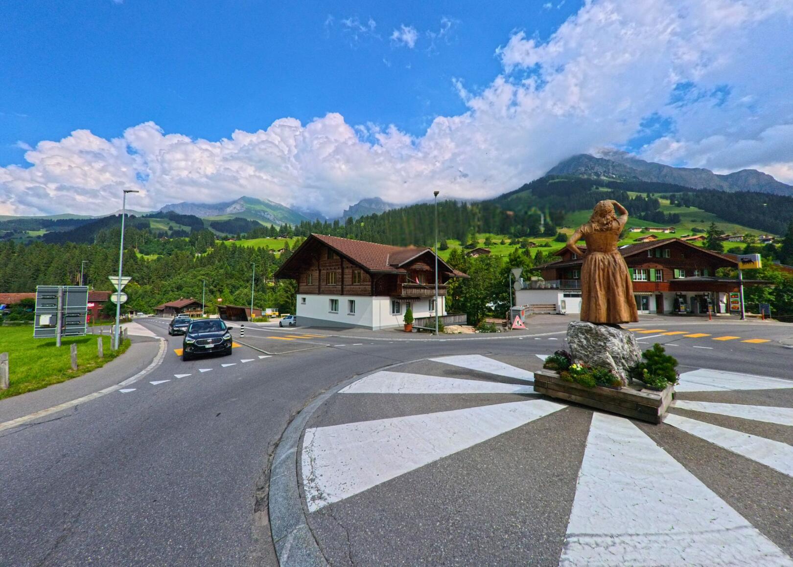 Im Kreisverkehr von Adelboden (3715, Kanton Bern) steht eine Holzskulptur mit Blick auf die umliegenden Berghänge und traditionellen Chalets vor bewaldeter Hügellandschaft.