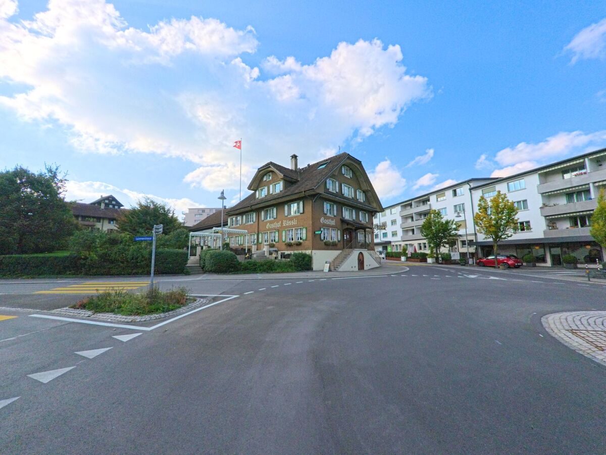 Im Zentrum von Adligenswil (6043, Kanton Luzern) steht das traditionelle Gasthaus Rössli mit Schweizer Fahne an einer Strassenkreuzung.