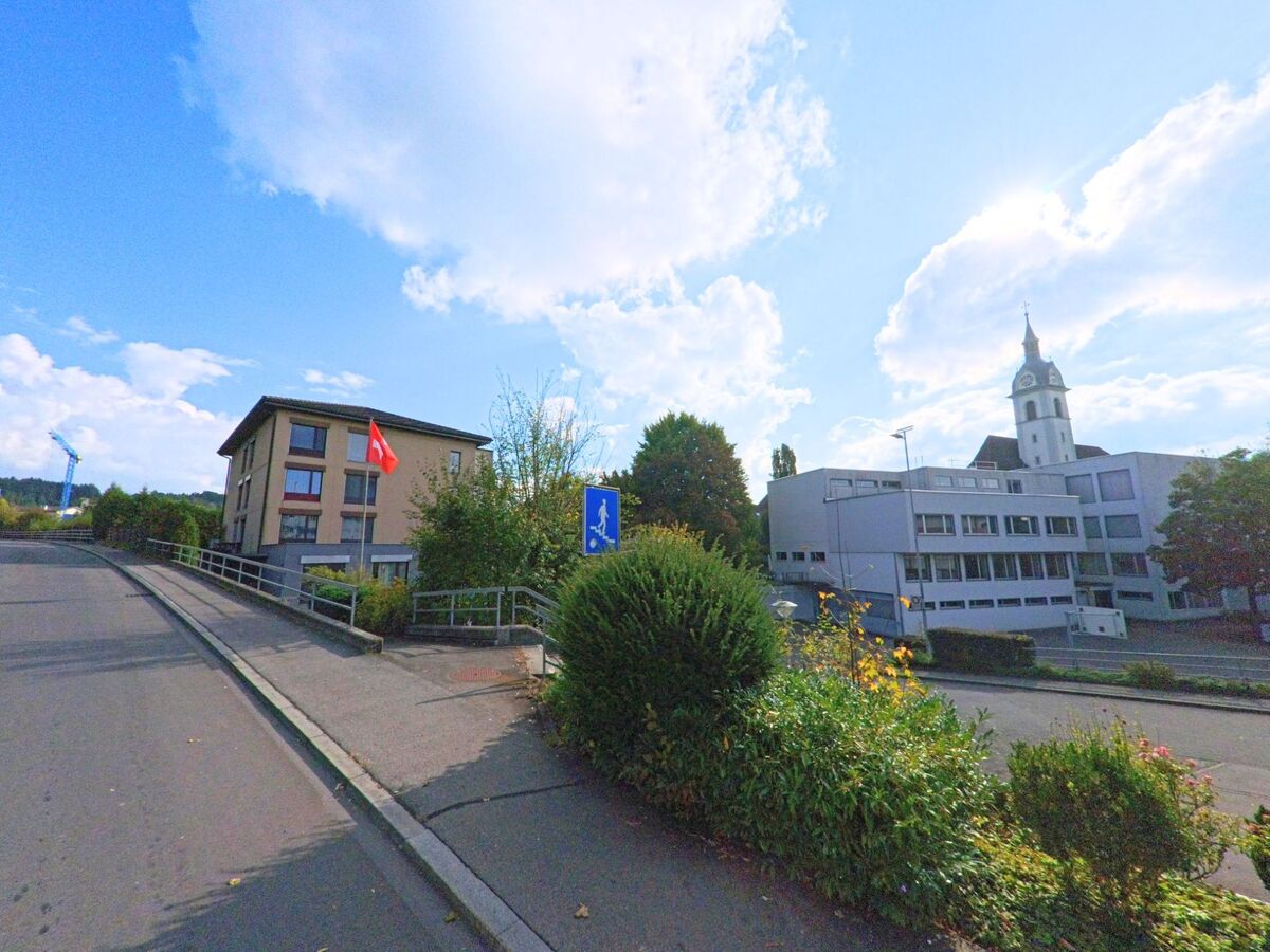 In Adligenswil (6043, Kanton Luzern) steht ein modernes Gebäude mit Schweizer Fahne nahe der Kirche und dem Gemeindezentrum.
