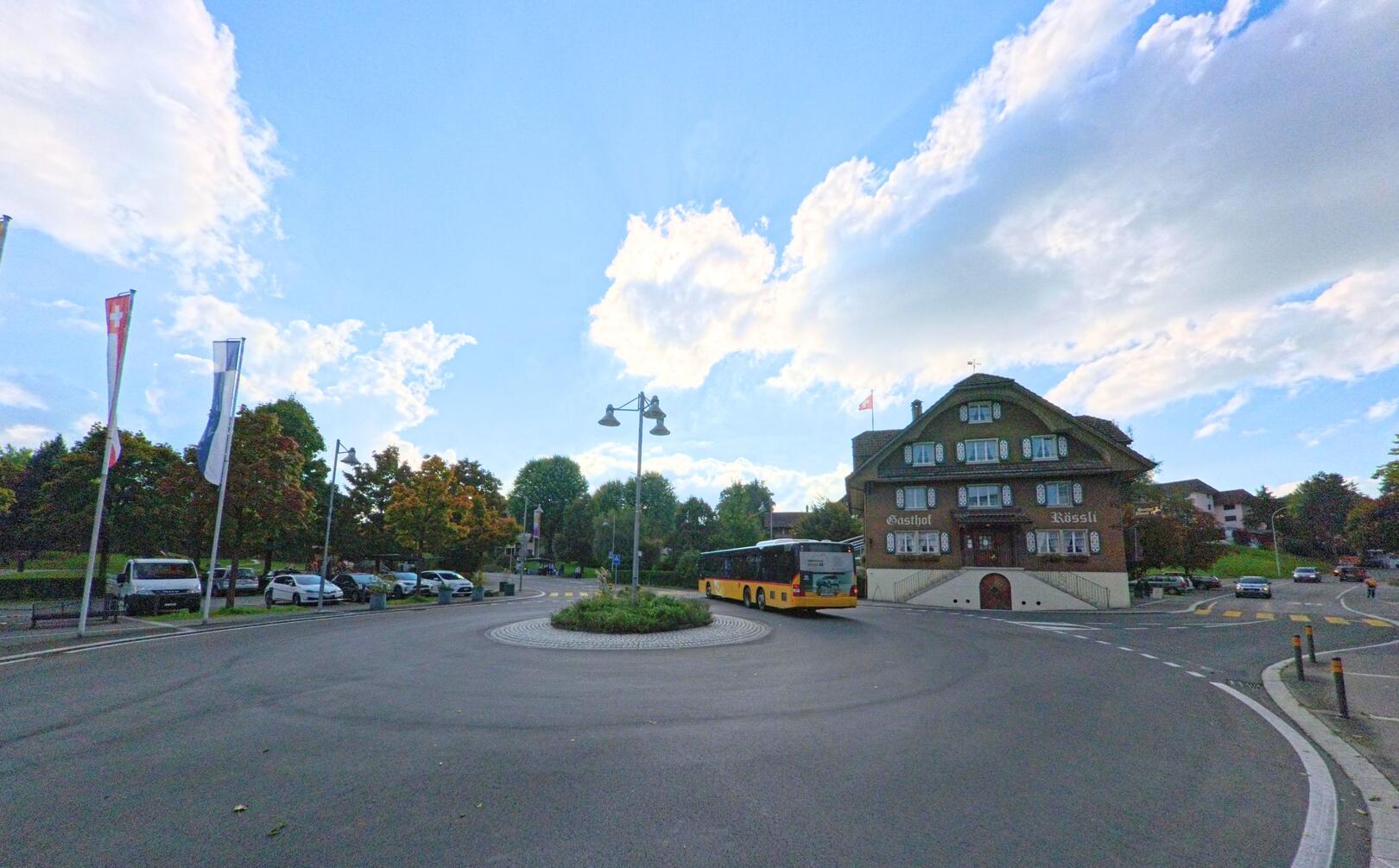 Ein Postauto fährt durch den Kreisverkehr in Adligenswil (6043, Kanton Luzern) vor dem historischen Gasthaus Rössli unter blauem Himmel.