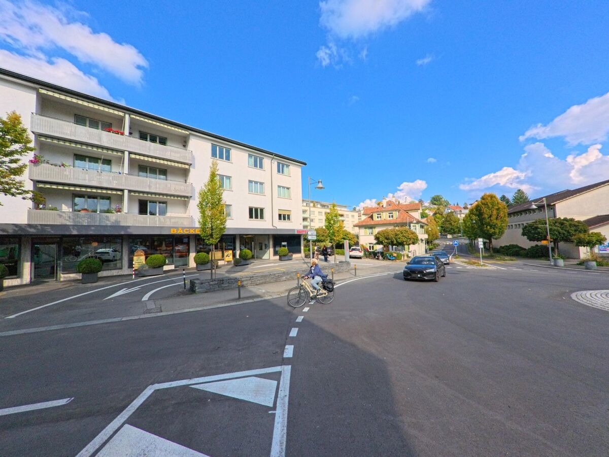 Eine belebte Kreuzung im Zentrum von Adligenswil (6043, Kanton Luzern) mit modernen Gebäuden, Geschäften und vorbeifahrenden Autos.