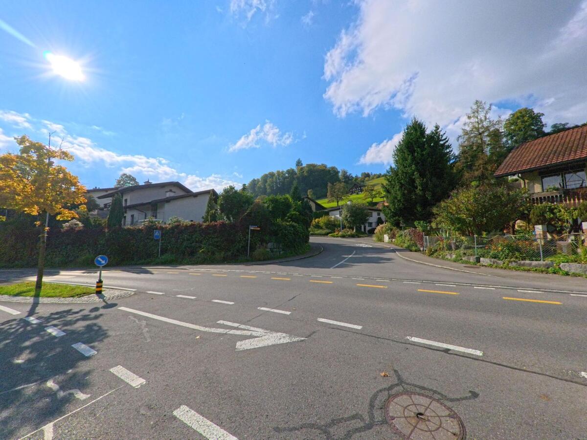 Eine sonnige Kreuzung in Adligenswil (6043, Kanton Luzern) führt zwischen Wohnhäusern in Richtung eines bewaldeten Hügels.
