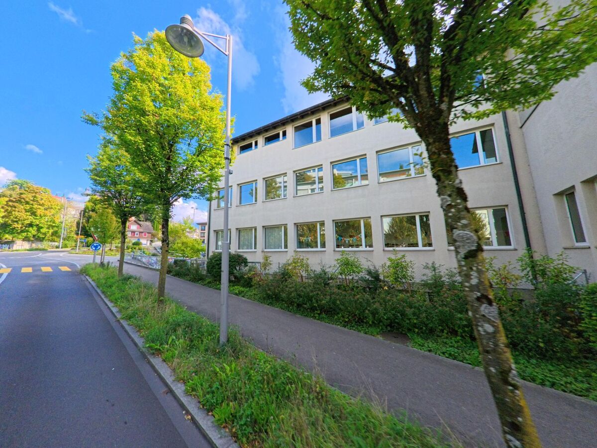 Ein modernes Schulgebäude in Adligenswil (6043, Kanton Luzern) liegt an einer ruhigen Strasse mit Bäumen und Gehweg.