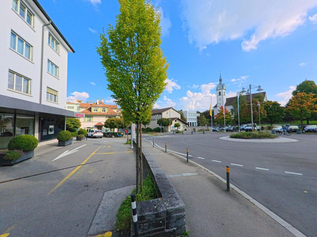 Im Zentrum von Adligenswil (6043, Kanton Luzern) steht die weisse Kirche mit Turm am Kreisverkehr, umgeben von modernen Gebäuden.
