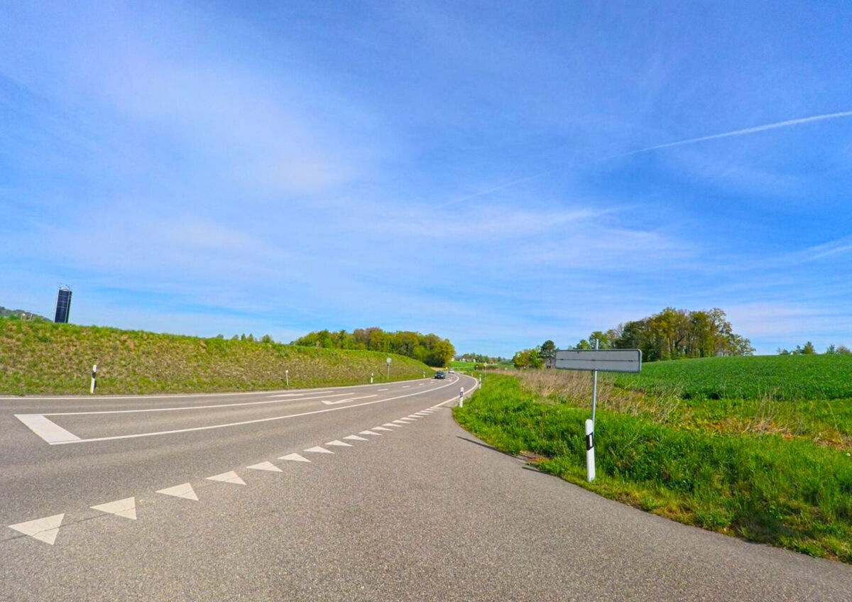 Eine Ausfallstrasse mit Markierungen und Blick auf grüne Felder verbindet Adlikon (8107, Kanton Zürich) mit der umliegenden Landschaft.