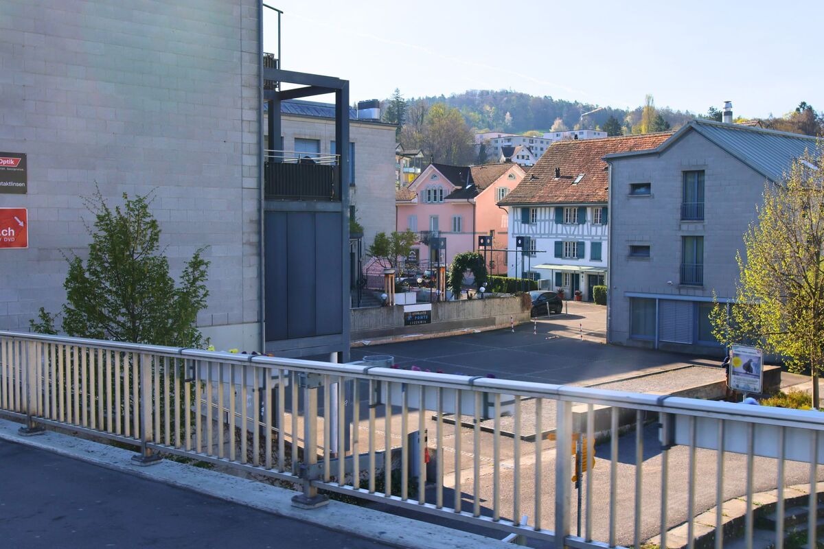  In Adliswil (8134, Kanton Zürich) bietet ein Blick von einer Brücke Sicht auf einen kleinen Platz mit traditionellen und modernen Gebäuden im Ortskern.