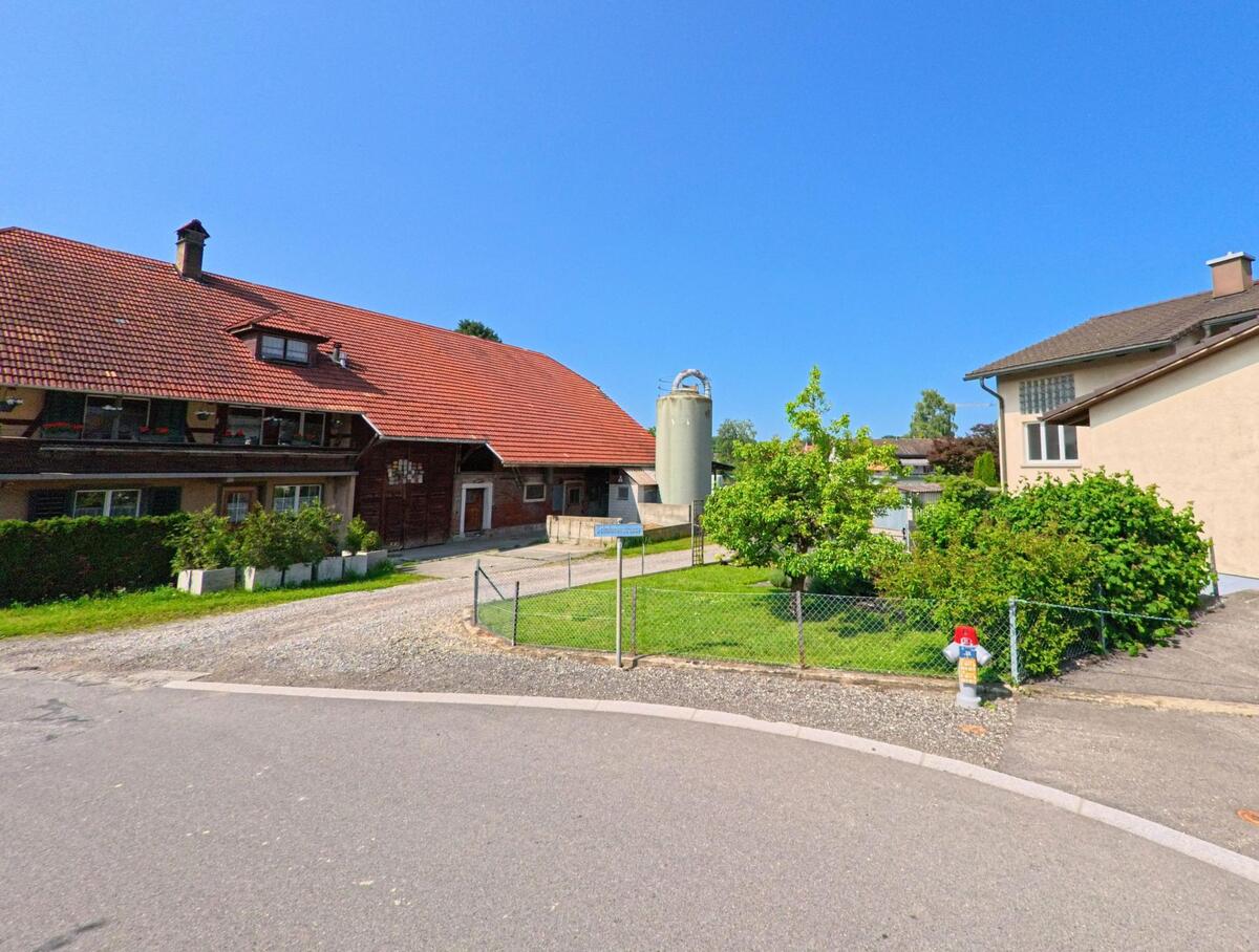 In Aefligen (3426, Kanton Bern) biegt die Straße an einem traditionellen Bauernhaus mit einem markanten roten Dach und einem großen Silos vorbei, während grünes Gras und ein Garten das Bild ergänzen.