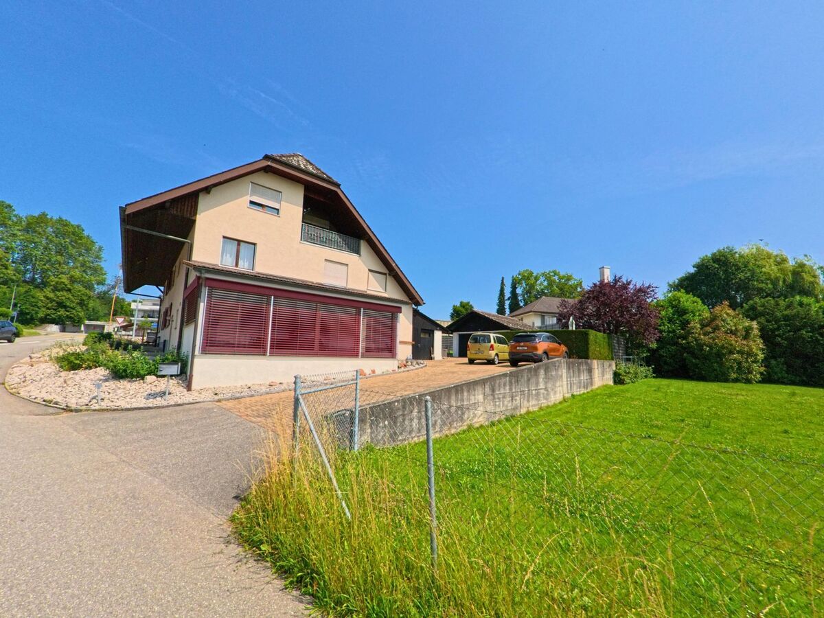 In Aeschi (4556, Kanton Solothurn) befindet sich ein Wohnhaus mit markanten Fensterlaeden an einer ruhig gelegenen Strasse, daneben eine Auffahrt mit geparkten Autos und angrenzender Gruenflaeche.