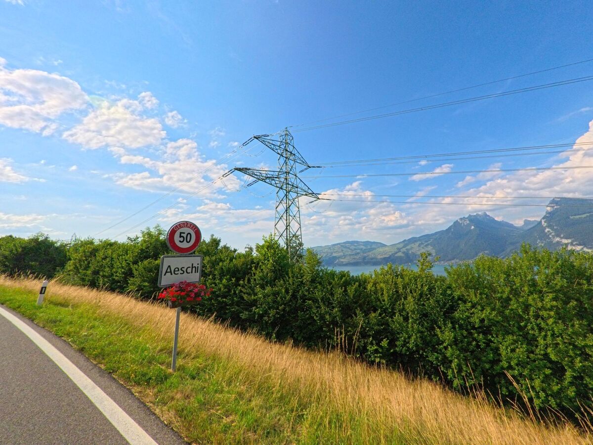 Das Ortsschild von Aeschi bei Spiez (3703, Kanton Bern) steht an einer Landstrasse mit Blick auf den Thunersee, eingerahmt von Stromleitungen und bewaldeten Hügeln.