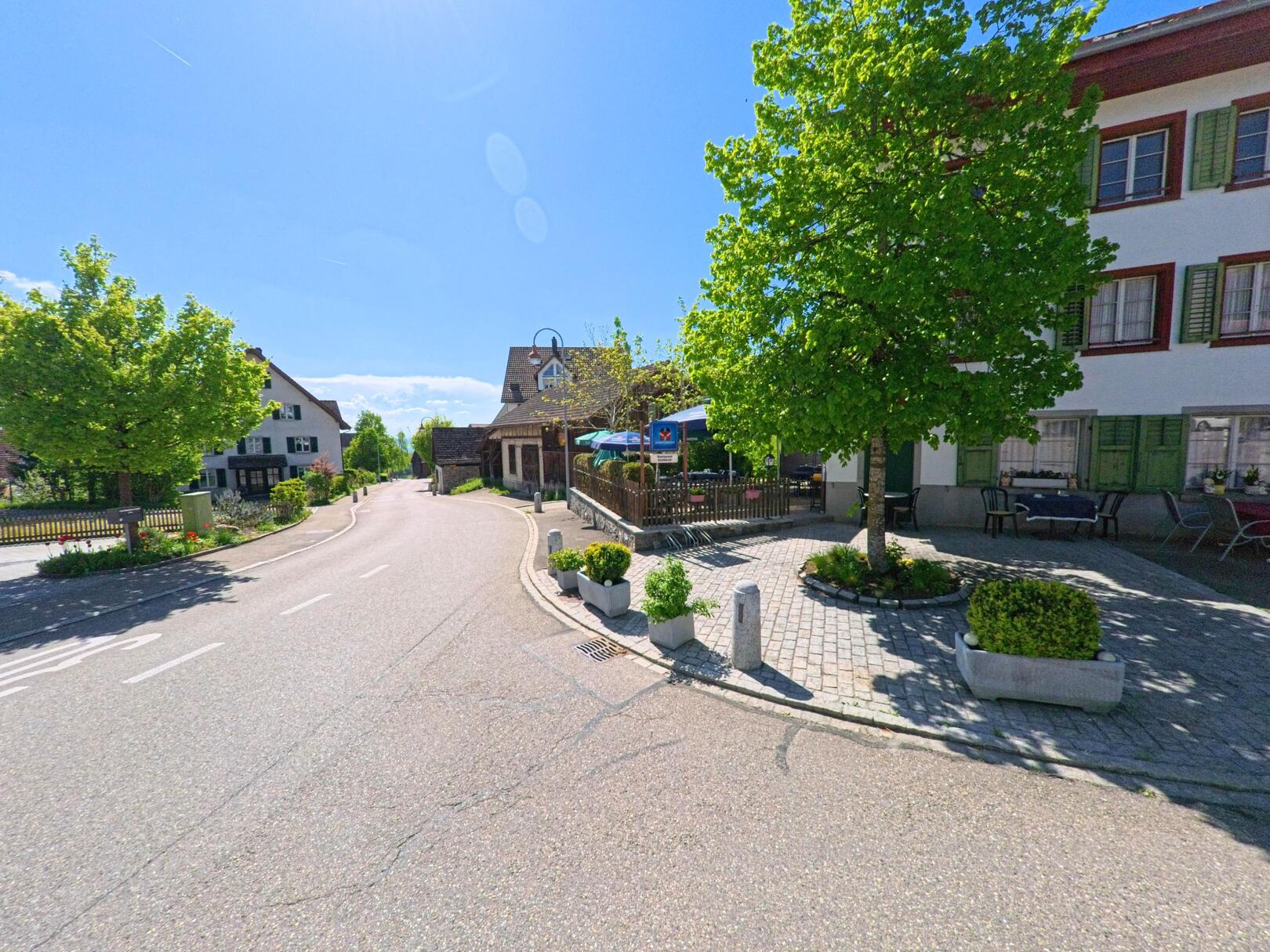 Im Zentrum von Aeugst am Albis, PLZ 8914 im Kanton Zürich, verläuft eine ruhige Strasse vorbei an traditionellen Häusern mit Fensterläden und einem Restaurant mit Gartenwirtschaft.