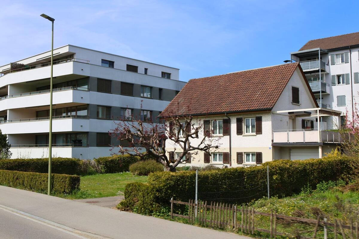 In Affoltern am Albis (PLZ 8909, Kanton Zürich) steht ein traditionelles Einfamilienhaus mit gepflegtem Vorgarten zwischen zwei modernen Mehrfamilienhäusern an einer ruhigen Strasse.