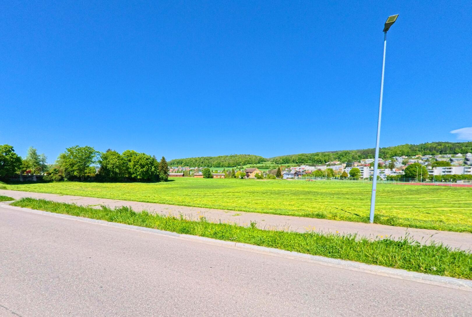 Am Rand von Affoltern am Albis (PLZ 8909, Kanton Zürich) verläuft ein Weg entlang einer weiten, frisch gemähten Wiese mit Blick auf die angrenzende Wohnsiedlung und bewaldete Hügel.