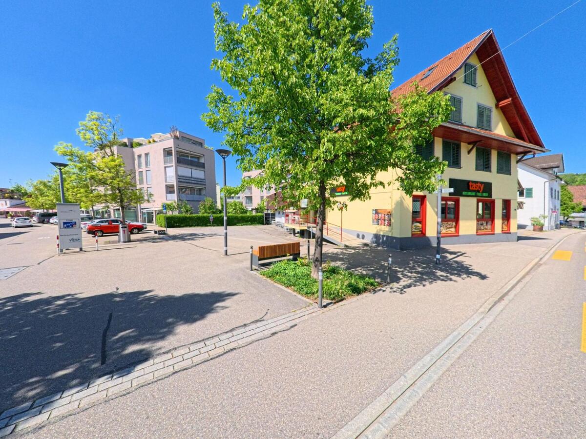 Im Zentrum von Affoltern am Albis (PLZ 8910, Kanton Zürich) steht ein gelb gestrichenes Gebäude mit Restaurantbetrieb, umgeben von Bäumen, Sitzbänken und moderner Wohnarchitektur.