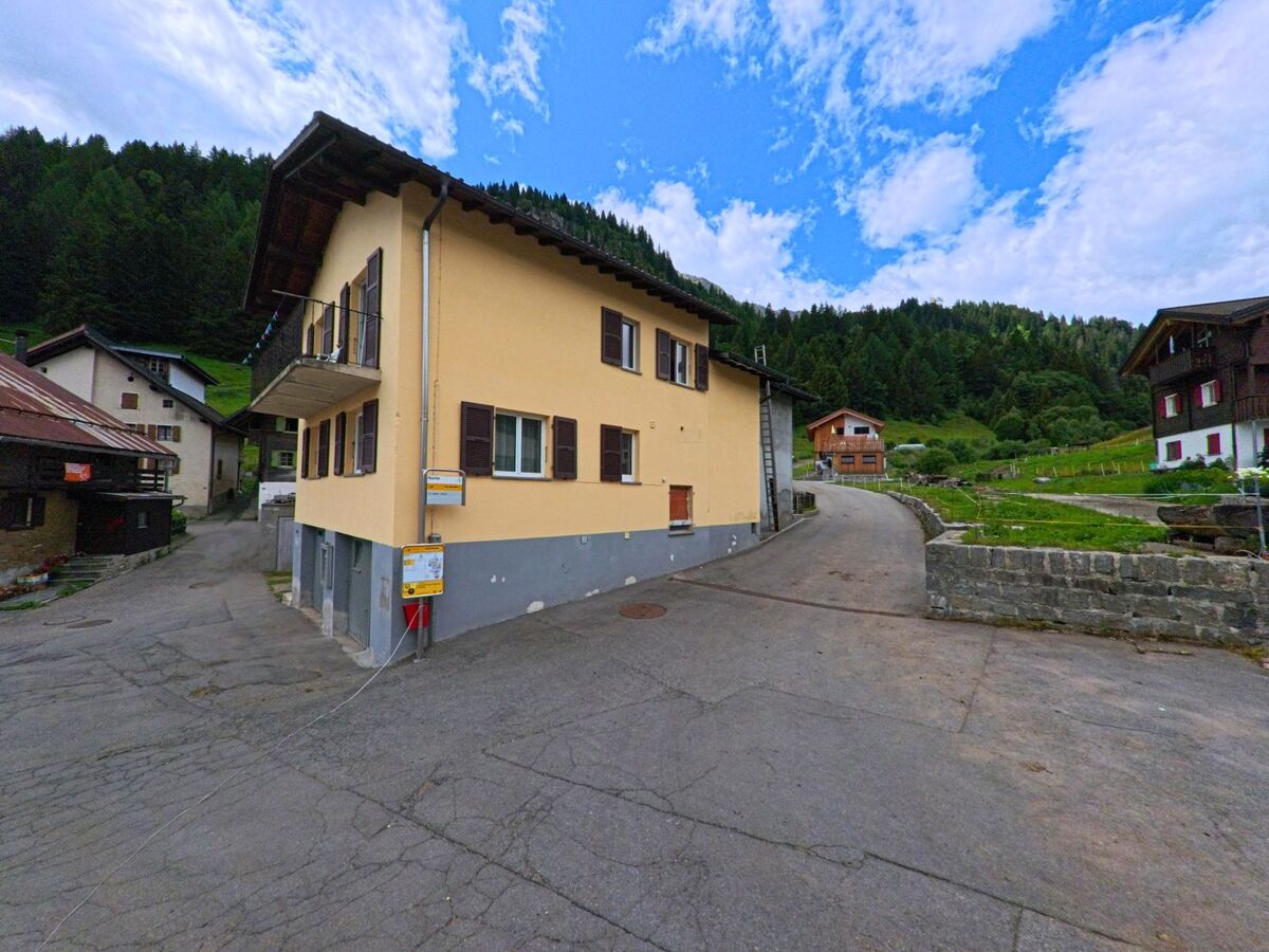 Ein gelbes Wohnhaus mit Bushaltestelle steht an einer schmalen Strasse in Airolo (6780, Kanton Tessin) mit Blick auf grüne Hänge und verstreute Holzhäuser.