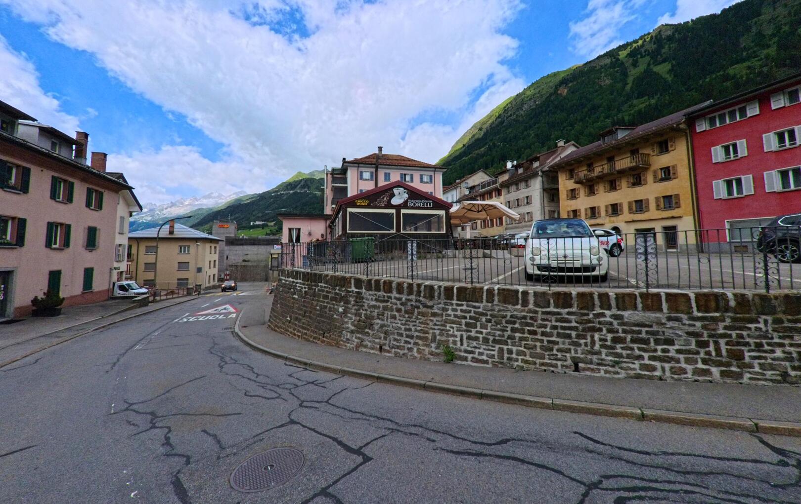 In Airolo (6780, Kanton Tessin) führt eine leicht ansteigende Strasse mit Wohnhäusern und dem Restaurant Borelli im Vordergrund in Richtung der umliegenden Berge.