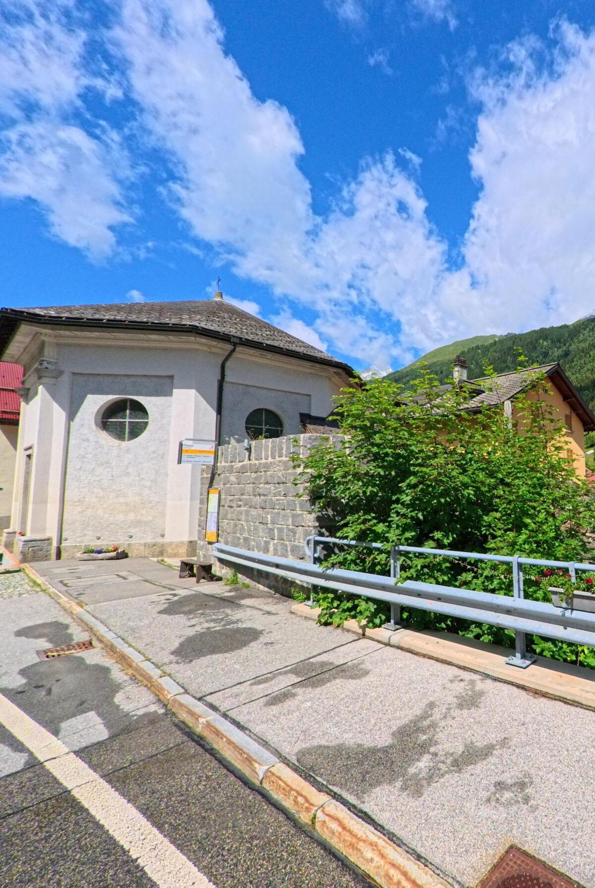 Eine kleine Kapelle mit rundem Fenster steht an einer Strasse in Airolo (6780, Kanton Tessin), umgeben von einer Steinmauer und grünen Büschen vor Bergkulisse.
