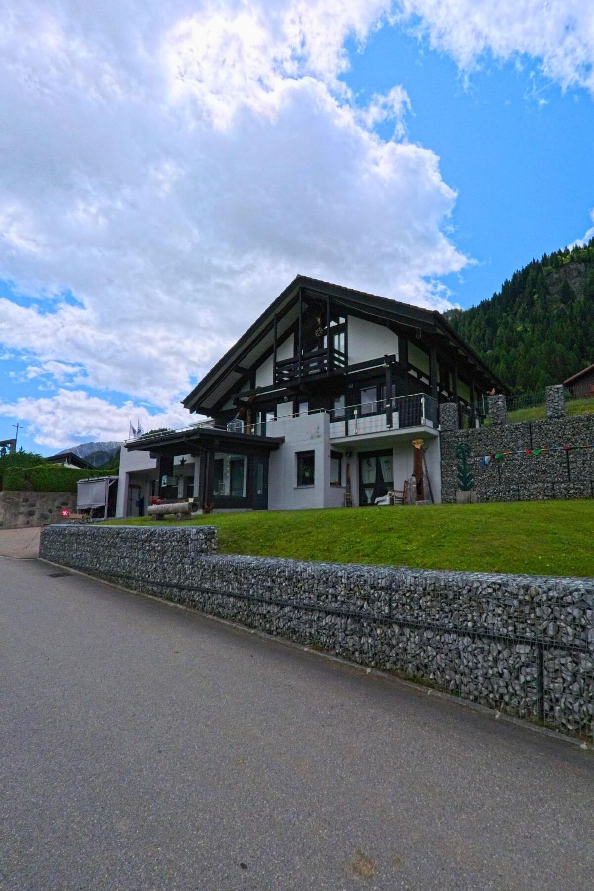 Ein modernes Chalet in Airolo (6780, Kanton Tessin) steht auf einer leicht ansteigenden Strasse, umgeben von grünen Hängen und Wald im Hintergrund.