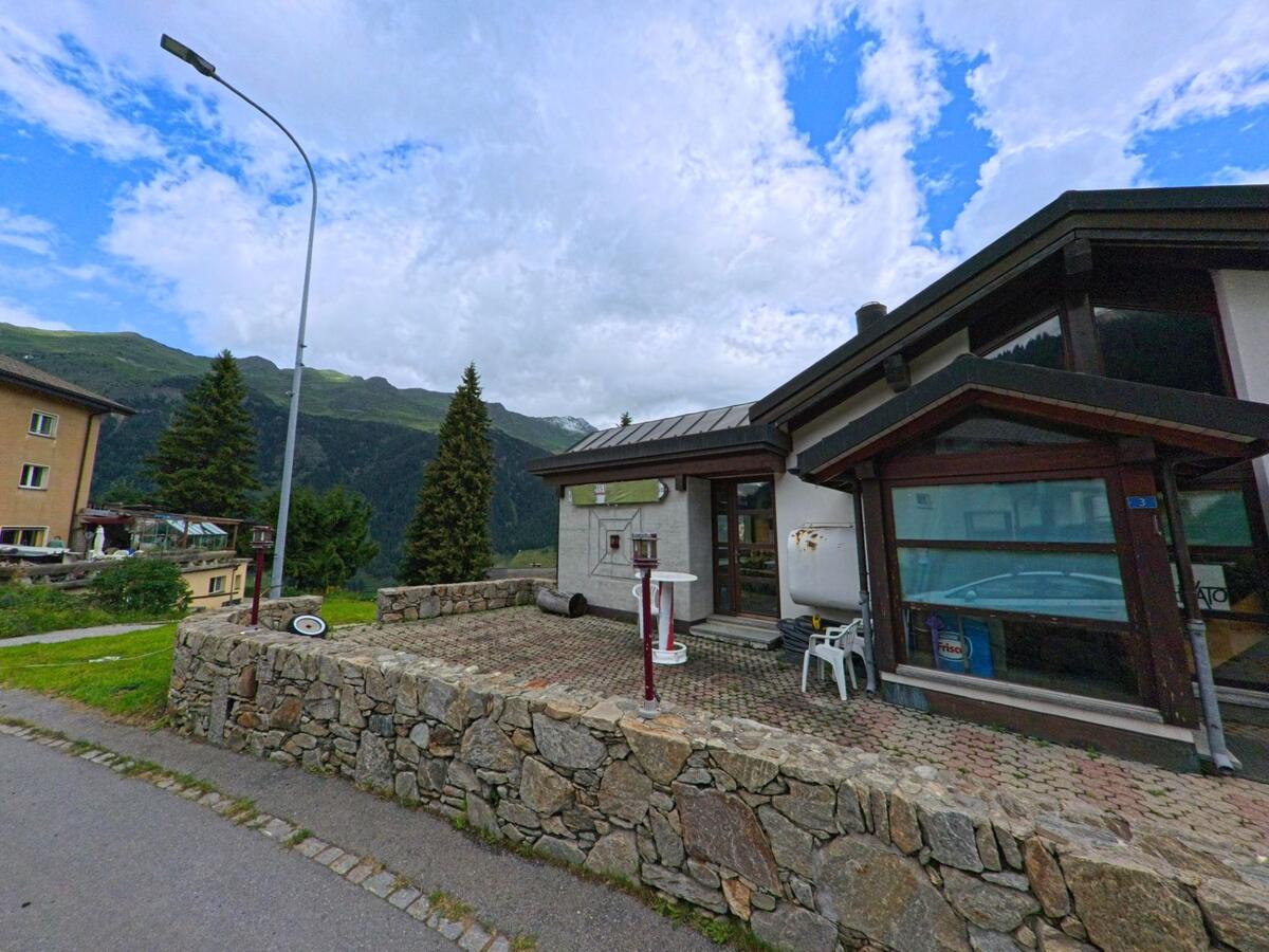 Ein modernes Haus mit grosser Glasfront und Natursteinmauer steht am Hang von Airolo (6780, Kanton Tessin) mit Blick auf bewaldete Berge im Hintergrund.