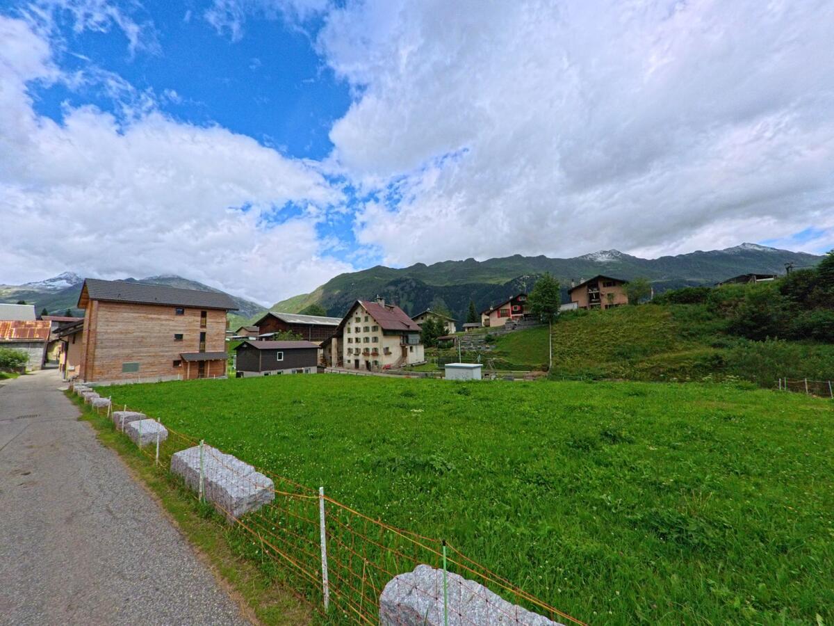 Zwischen grünen Wiesen und traditionellen Häusern liegt ein ruhiger Ortsteil von Airolo (6780, Kanton Tessin) mit Blick auf bewaldete Berge und schneebedeckte Gipfel.