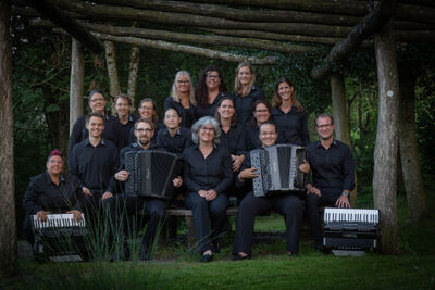 Unter einer hölzernen Pergola in Kriens (6010, Kanton Luzern) sitzt und steht das Akkordeon-Orchester Kriens in dunkler Kleidung fuers Gruppenfoto zusammen.