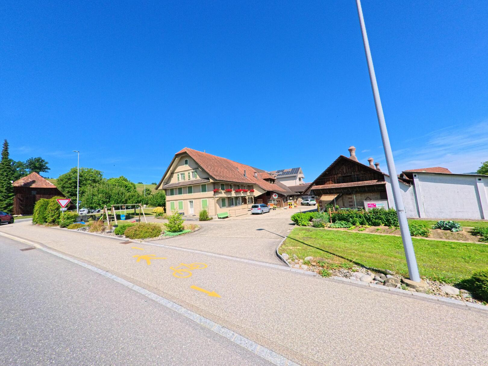 In Alberswil (6248, Kanton Luzern) steht ein stattlicher Bauernhof mit gruener Fensterlaeden und Blumenschmuck an einem Radweg mit gelber Markierung, umgeben von Gartenflaechen und Nebengebaeuden.