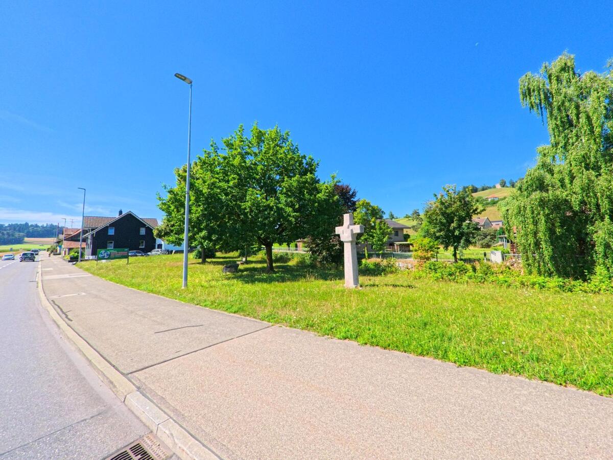 In Alberswil (6248, Kanton Luzern) steht ein steinernes Kreuz auf einer Gruenflaeche am Strassenrand, umgeben von Baeumen und Haeusern mit Blick auf die Huegellandschaft.