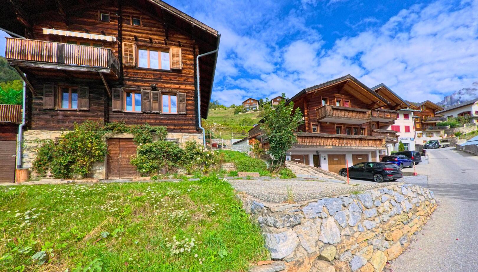 In Albinen (3955, Kanton Wallis) stehen gepflegte Chalets aus Holz entlang einer ansteigenden Strasse, umgeben von grünen Hängen und blauem Himmel.