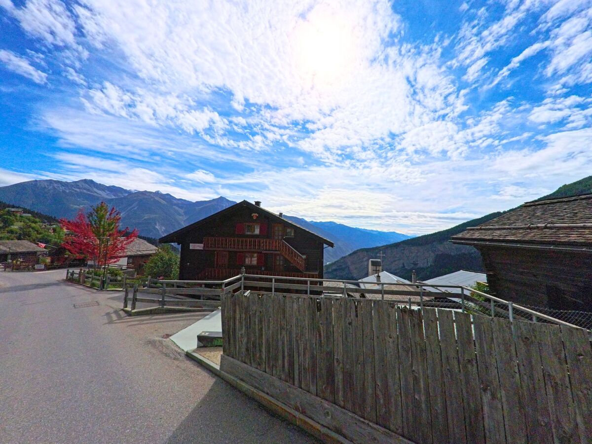 Ein traditionelles Holzhaus mit roten Fensterläden in Albinen (3955, Kanton Wallis) steht vor einer Bergkulisse, nebenan leuchtet ein Baum in Herbstfarben.