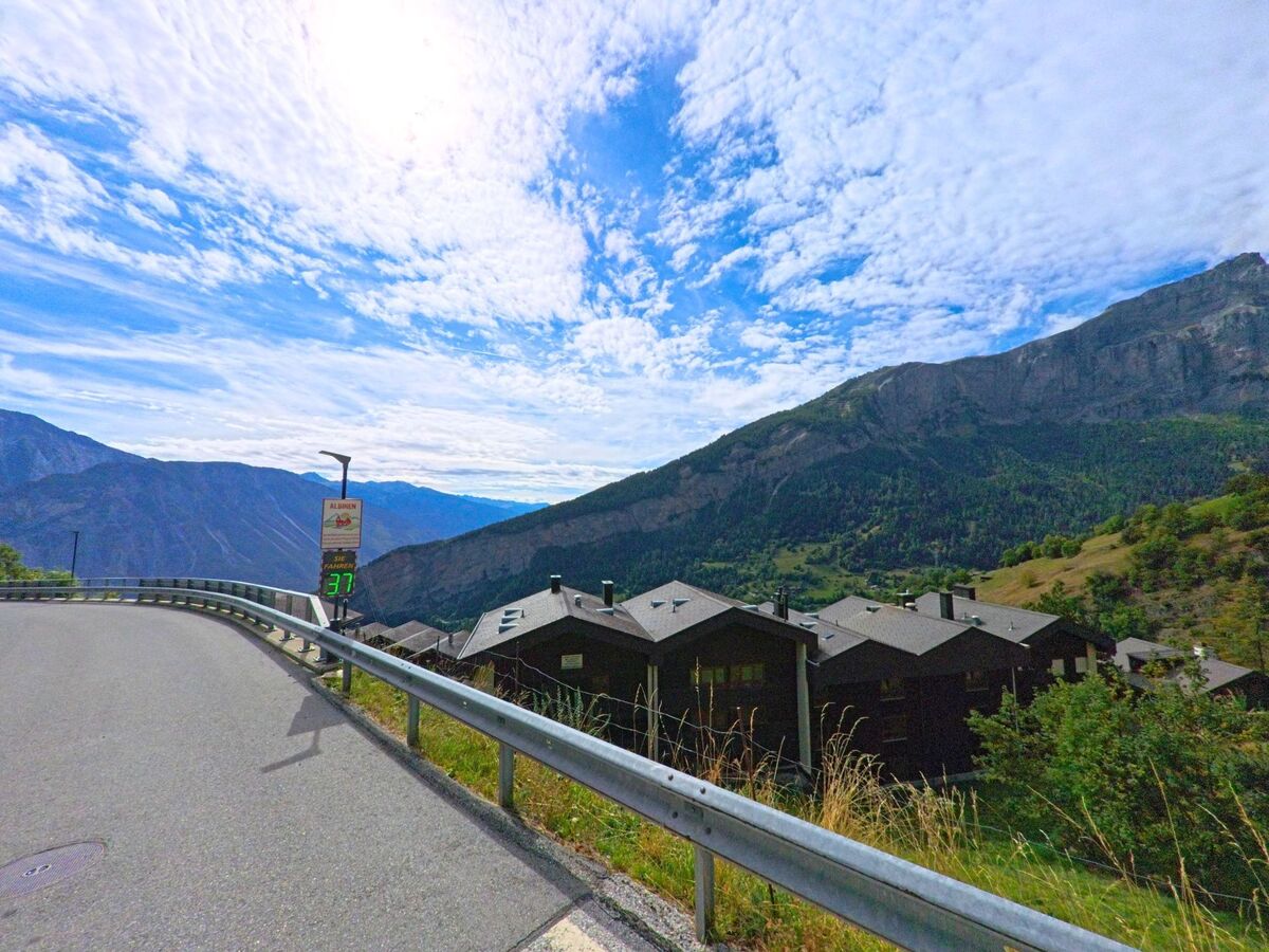 Eine kurvige Strasse führt oberhalb von Albinen (3955, Kanton Wallis) entlang, mit Blick auf dunkle Häuserdächer und die umliegenden Berge.