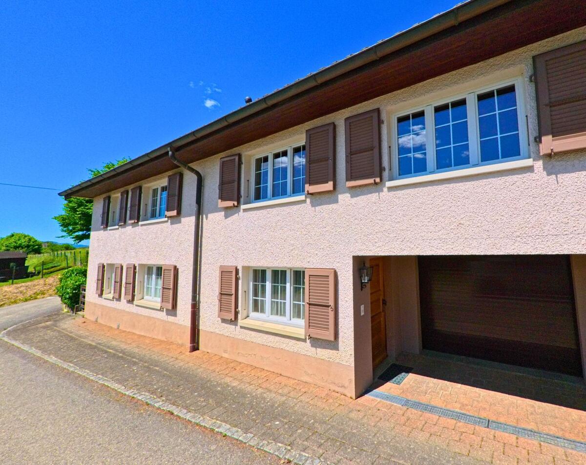 Ein langgezogenes Wohnhaus mit braunen Fensterläden steht in Alle (2942, Kanton Jura) an einer schmalen Strasse mit leichtem Kurvenverlauf.